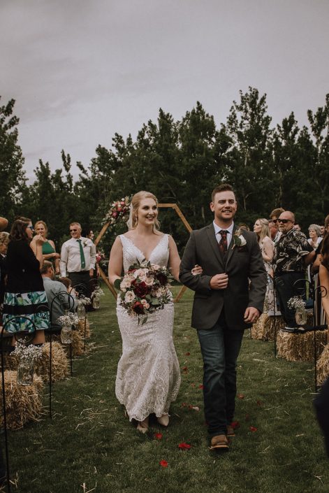 Bride and groom walking from ceremony holding rustic red and blush bridal bouquet featuring red charm peonies, quicksand roses, blush spray roses, burgundy astrantia, light pink astilbe and eucalyptus greenery.