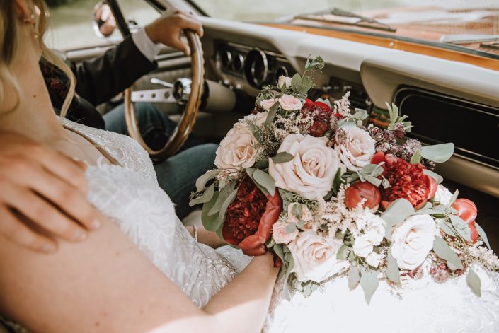Bride and groom in an antique car holding a rustic red and blush bridal bouquet featuring red charm peonies, quicksand roses, blush spray roses, burgundy astrantia, and light pink astilbe with eucalyptus greenery.