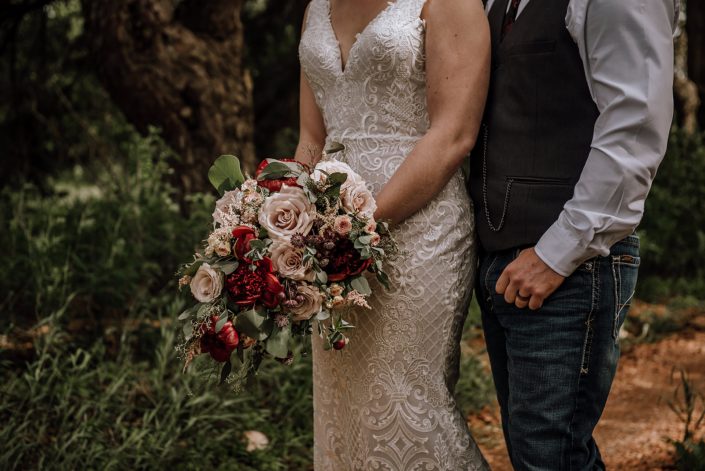 Bride and groom standing together with a rustic red and blush bridal bouquet made of red charm peony, quicksand roses, blush spray roses, light pink astilbe, burgundy astrantia and eucalyptus greenery.