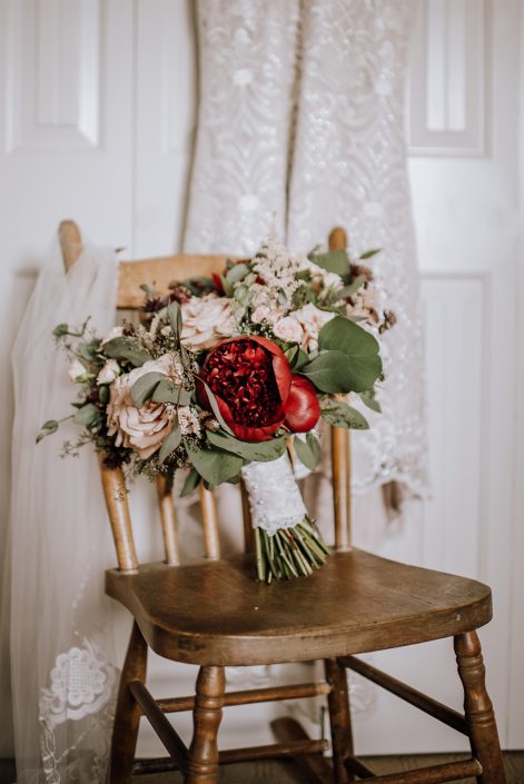 Bride's bouquet, veil and dress with a vintage rustic wooden chair. Bouquet features red charm peony, quicksand roses, burgundy astrantia, pale pink astilbe, and blush spray roses. It was finished with eucalyptus greenery and a blush satin with lace overlay wrapped handle.