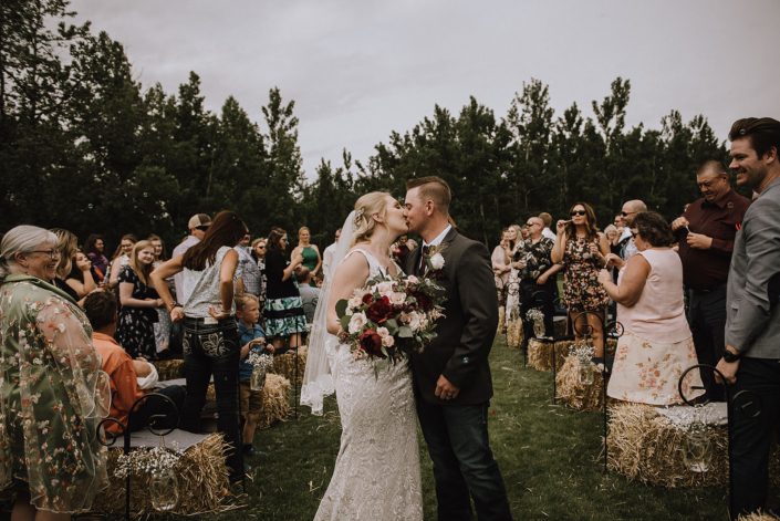 Bride and groom kissing at the ceremony; bride holding rustic red and blush bouquet featuring red charm peonies, blush quicksand roses and eucalyptus greenery.
