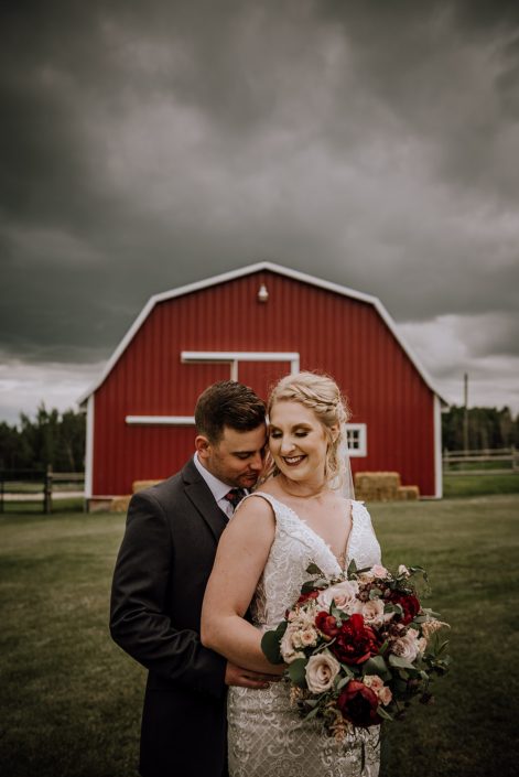 Bride and groom, Courtney and Caleb, in front of a red barn with grey clouds overhead; rustic red and blush bridal bouquet designed with red charm peonies, quicksand roses, burgundy astrantia, pale pink astilbe, and blush spray roses with eucalyptus greenery.
