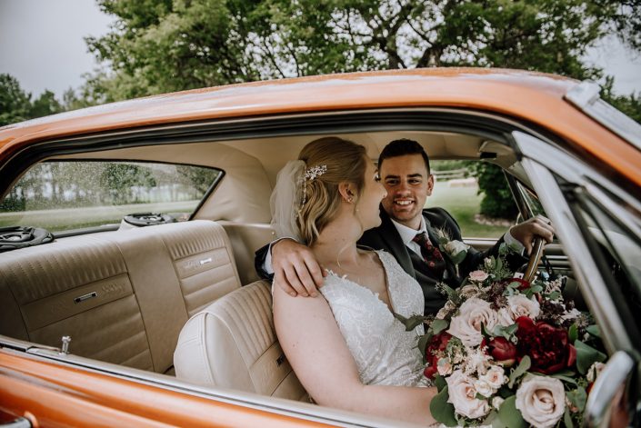 Bride and groom looking at each other sitting in an antique car; bride holding a rustic red and blush bridal bouquet designed with red charm peonies, quicksand roses, burgundy astrantia, light pink astilbe, and blush spray roses with eucalyptus greenery.