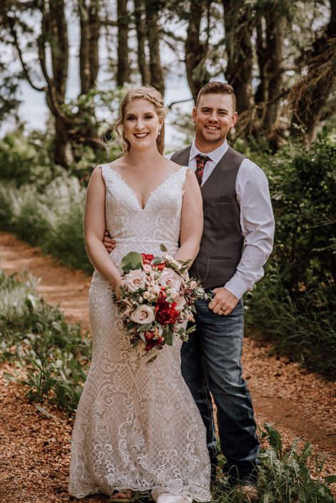 Groom wearing a grey vest and jeans standing with bride wearing a white lace gown and holding a rustic red and blush bridal bouquet featuring red charm peonies, quicksand roses, blush spray roses, burgundy astrantia, light pink astilbe and eucalyptus greenery.