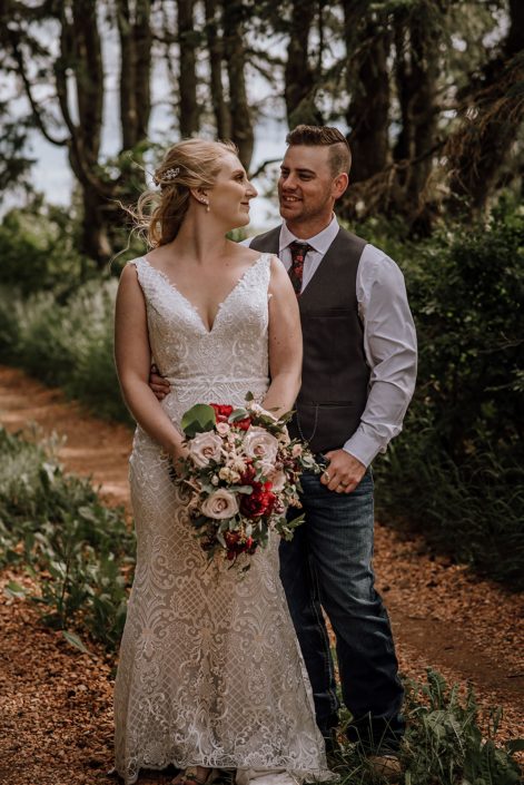 Bride and groom looking at one another; bride holding a rustic red and blush cascading bouquet designed with red charm peonies, quicksand roses, blush spray roses, burgundy astrantia, pale pink astilbe, and eucalyptus greenery.