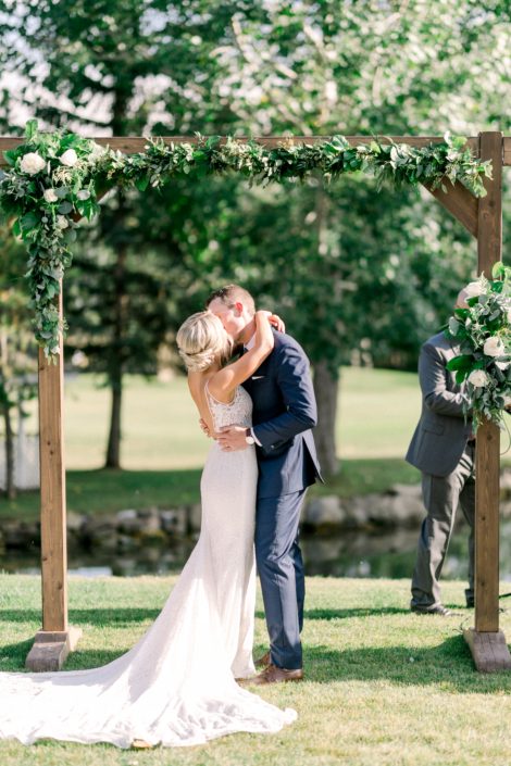 Kayla and Joel kissing under a wooden archway covered with arrangements and garlands made of salal, Italian ruscus, plumosa, eucalyptus and Tibet roses.