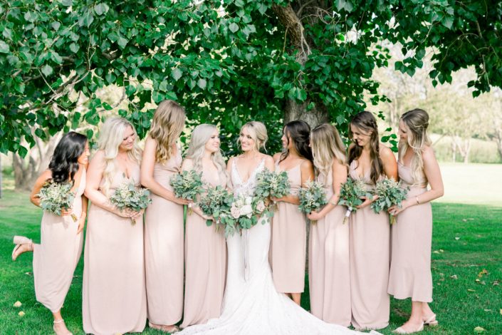 Kayla wearing a white lace dress and holding a white and blush bouquet surrounded by her bridesmaids wearing blush floor-length dresses and holding bouquets made of white astilbe, olive branches and eucalyptus greenery.