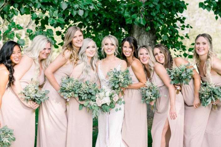 Kayla wearing a white bridal gown and holding a bridal bouquet featuring white o'hara garden roses, white ranunculus, quicksand roses, white astilbe, olive branches and eucalyptus surrounded by her bridesmaids wearing blush floor-length dresses and holding simple bouquets made of white astilbe, olive branches and eucalyptus greenery.