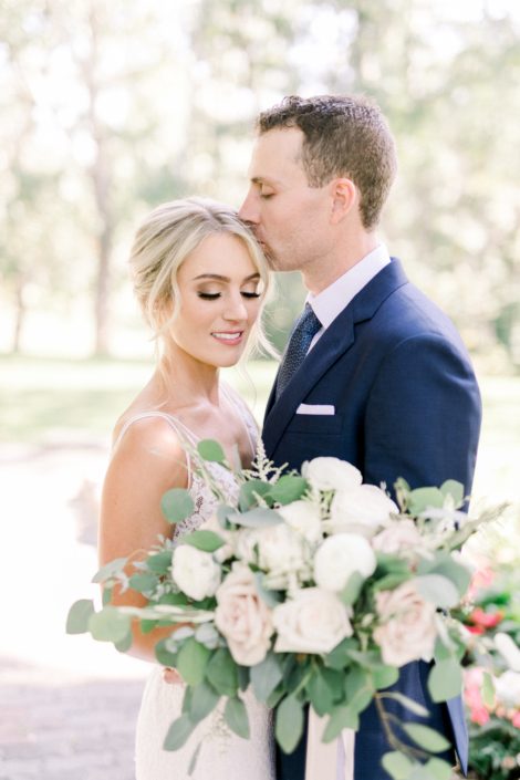 Joel kissing bride Kayla holding a blush and white bouquet featuring white o'hara garden roses, white ranunculus, quicksand roses, white astilbe, olive branches and fresh eucalyptus greenery.