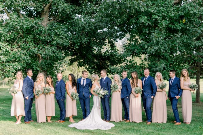 Timeless white wedding bridal party; bride wearing white lace bridal gown and holding a blush and white bouquet; bridesmaids wearing blush floor-length gowns and holding simple white astilbe and greenery bouquets; groom and groomsmen wearing navy suits.