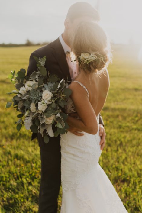 Bride and groom, Leah and Chance; bride hair flowers designed with white spray roses, lisianthus and astilbe with a touch of greenery; bridal bouquet featuring white Playa Blanca roses, white lisianthus, white astilbe, Blue Star succulents, dusty miller and eucalyptus greenery.