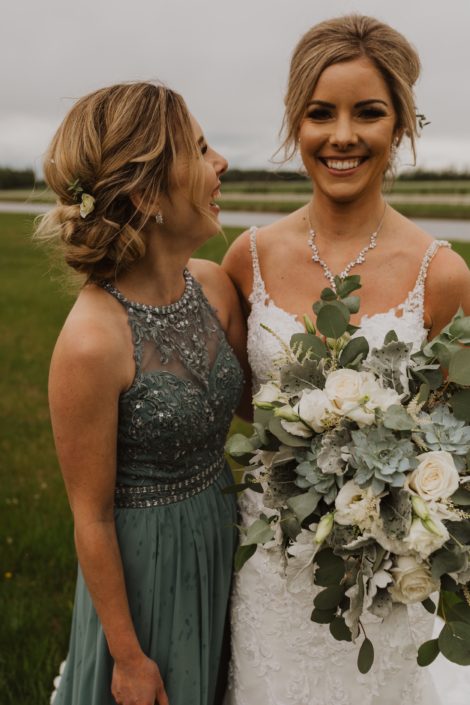 Bride wearing white lace dress and holding a vintage inspired white and grey green bridal bouquet featuring playa blanca roses, lisianthus, astilbe, Blue Star succulents, dusty miller and eucalyptus; bridesmaid wearing grey green bridesmaid dress with white hair flowers.