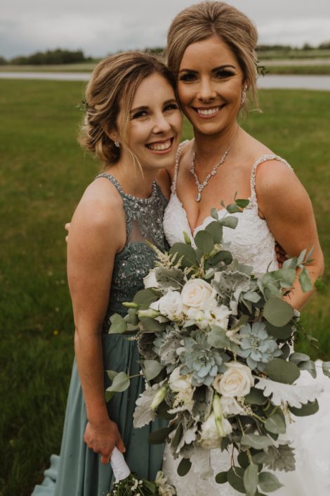 Bride and bridesmaid; bride wearing a white lace dress and holding a vintage inspired white and grey green bridal bouquet designed with Playa Blanca roses, lisianthus, astilbe, blue star succulents, dusty miller and a mixed variety of eucalyptus; bridesmaid is wearing a grey green dress.