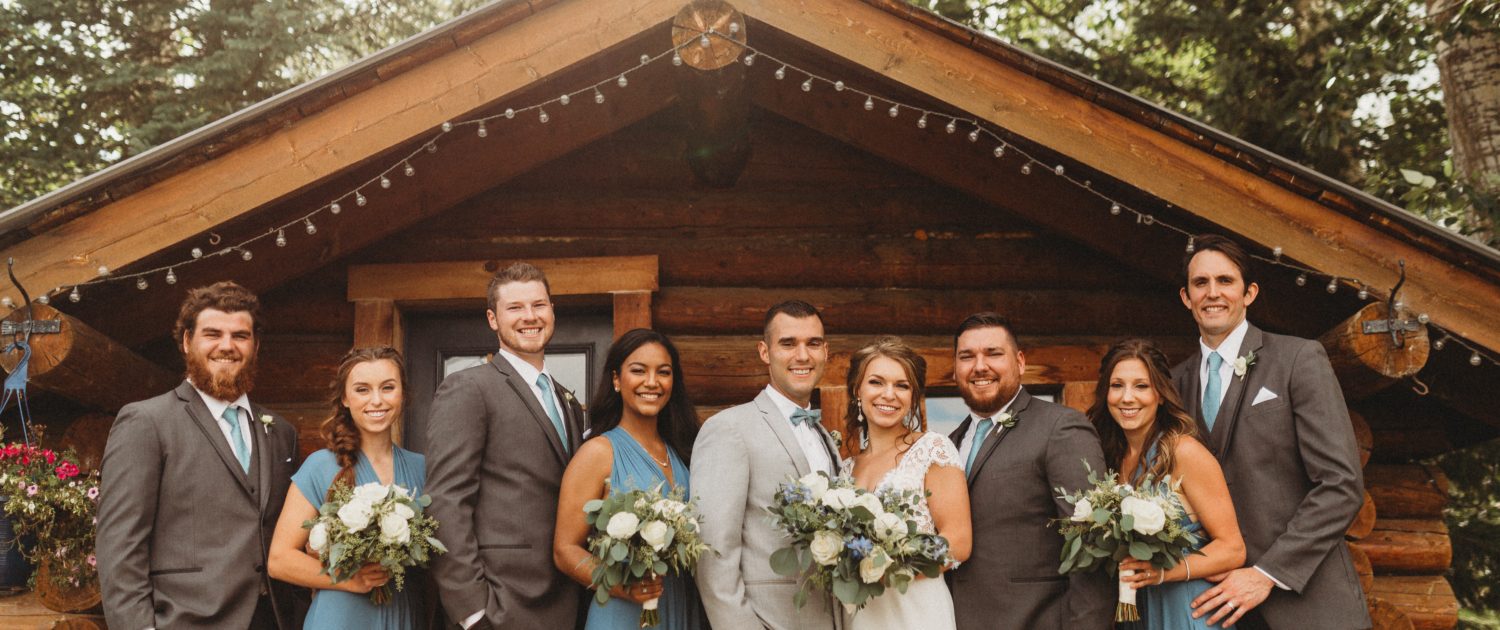 Kaitlin and Carter's blue and white bridal party in front of Cabin at Pine and Pond; bride and bridesmaids holding blue and white bouquets featuring roses and ranunculus; groom and groomsmen wearing white and blue boutonnieres.