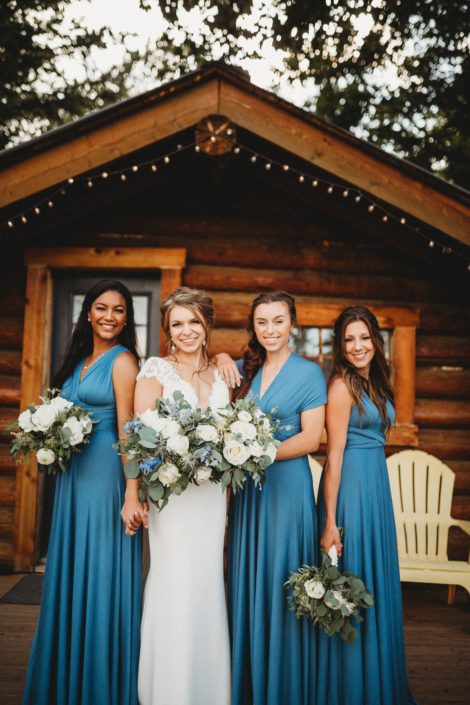 Bride and bridesmaids in front of a cabin at Pine and Pond; bridesmaid wearing white lace gown and holding blue and white bouquet; bridesmaids wearing blue and holding bouquets designed with white flowers and eucalyptus greenery.