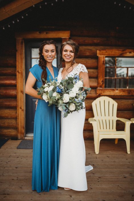 Bride holding blue and white bouquet standing next to bridesmaid wearing a dusty blue floor-length gown and holding a bouquet designed with white roses, ranunculus, astilbe and eucalyptus greenery.
