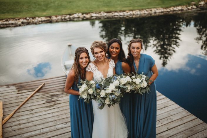 Kaitlin and Carter's Blue and White Pine & Pond Wedding - Bride and bridesmaids in front of pond at Pine and Pond with blue and white bouquets designed with roses, ranunculus, delphinium, eryngium and astilbe with eucalyptus greenery.