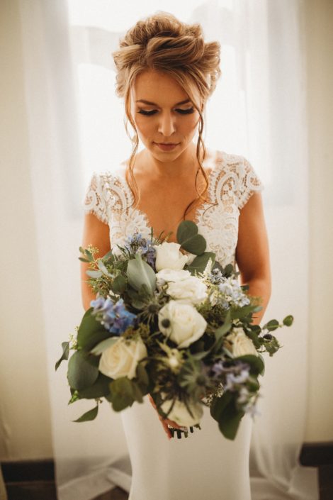 Bride, Kaitlin, holding dusty blue and white bouquet designed with Tibet roses, white ranunculus, astilbe, blue delphinium, blue eryngium and grey toned eucalyptus greenery.