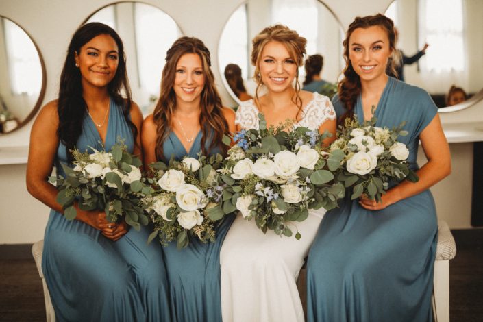 Bride, Kaitlin and her bridesmaids; bridal bouquet designed with Tibet roses, white ranunculus, white astilbe, blue delphinium, dusty blue eryngium, and eucalyptus greenery; bridesmaids bouquets made of similar white flowers.