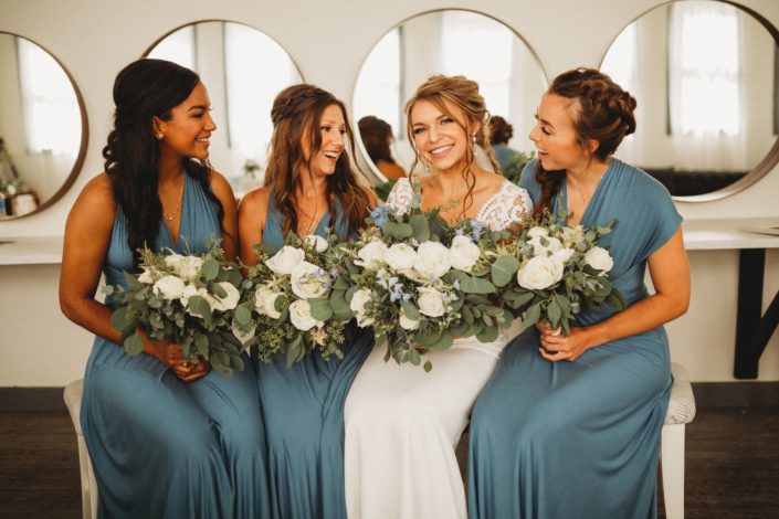 Bride and bridesmaids wearing dusty blue and holding bouquets designed with Tibet roses, white ranunculus, white astilbe, delphiniums, eryngium and eucalyptus.