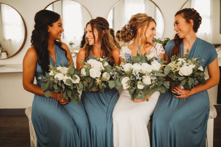 Bride and her bridesmaids holding white and dusty blue bouquets made of roses, ranunculus, astilbe, eryngium, delphinium, and eucalyptus.