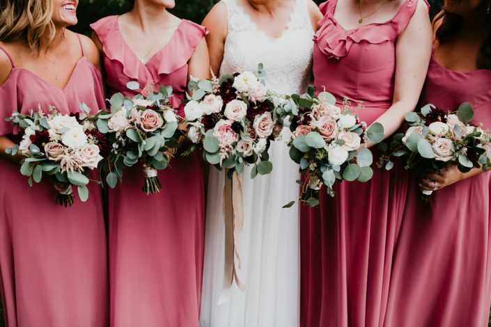 Bride standing alongside bridesmaids wearing pink berry floor length dresses and holding bouquets designed with burgundy dahlias, white ranunculus, roses, astilbe and eucalyptus.
