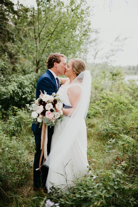 Bride kissing groom and holding a jewel tone bridal bouquet designed with burgundy dahlias, keira garden roses, white ranunculus, amnesia roses, quicksand roses, blackberry scoop scabiosa, light pink astilbe and eucalyptus greenery.
