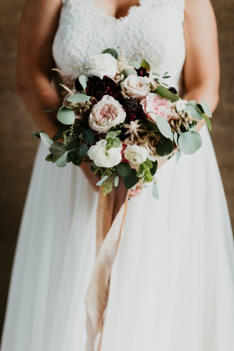 Bridal bouquet featuring burgundy dahlias, Keira garden roses, white ranunculus, amnesia roses, quicksand roses, blackberry scoop scabiosa, light pink astilbe and a mixed variety of eucalyptus greenery finished with trailing ribbons.