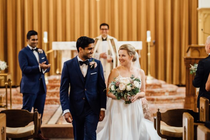 Bride and groom walking up aisle; groom wearing boutonniere; bride holding blush bouquet made with quicksand roses, white o'hara garden roses, white ranunculus, light pink astilbe and a variety of eucalyptus greenery.