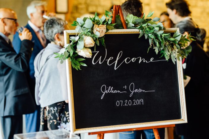 Jill and Jason's "Welcome" sign decorated with a fresh greenery garland made of eucalyptus and italian ruscus accented by quicksand roses.