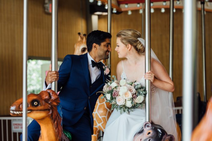Bride and groom, Jill and Jason, on a carousel at the Calgary Zoo; bride holding romantic blush bouquet featuring white o'hara garden roses, white ranunculus, quicksand roses, light pink astilbe and eucalyptus greenery.