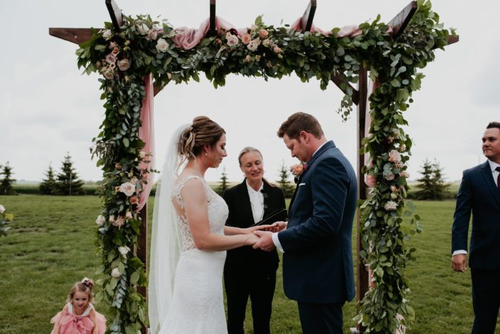 Crystal and Chance during their wedding ceremony under a wooden archway covered with a fresh greenery garland with accents of fresh blooms including burgundy dahlias, quicksand roses and cappuccino roses.