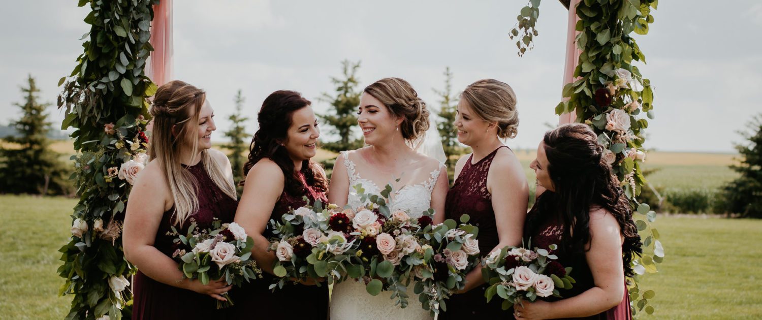 Crystal's bridesmaids standing under a wooden archway covered in a fresh greenery garland adorned with flowers including burgundy dahlias, cappuccino roses, quicksand roses, and astilbe.