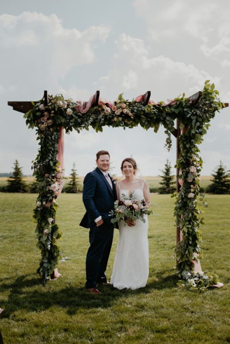 Crystal and Chance standing under wooden archway covered with a fresh greenery garland made of salal and eucalyptus accented by a variety of fresh blooms including dahlias, astilbe, quicksand roses and cappuccino roses.