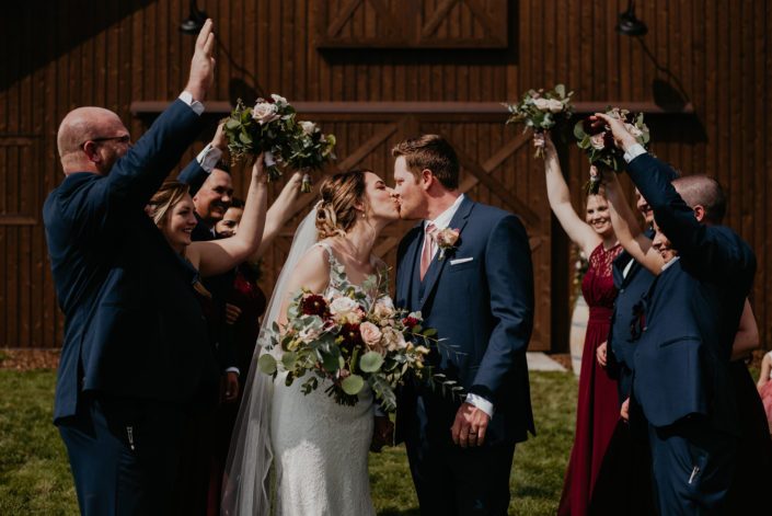 Bride and groom kissing surrounded by cheering bridal party; groom wearing boutonniere made of spray roses, sweet pea and astilbe; bride holding bridal bouquet featuring burgundy dahlias, burgundy cymbidium, astrantia, peach ranunculus, cappuccino roses, quicksand roses and a variety of eucalyptus greenery.