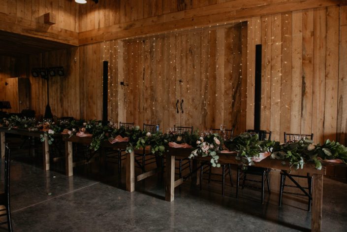 Head table covered in a fresh greenery garland with barn wall backdrop and twinkly lights.
