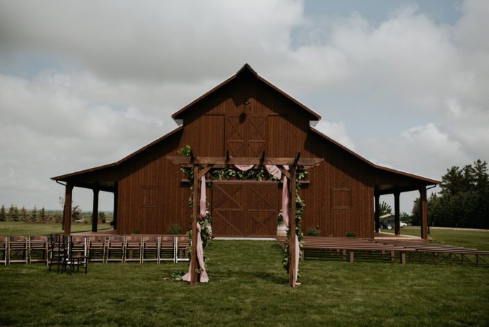 Sweet haven barn ceremony set up including wooden pews and a wooden archway covered in a fresh greenery garland with burgundy and dusty rose flowers.