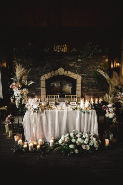 Boho Glam sweetheart table in front of a grand stone fireplace decorated with floral arrangements, fresh loose greenery, ivory linens, white pillar candles, and gold geometric accents.