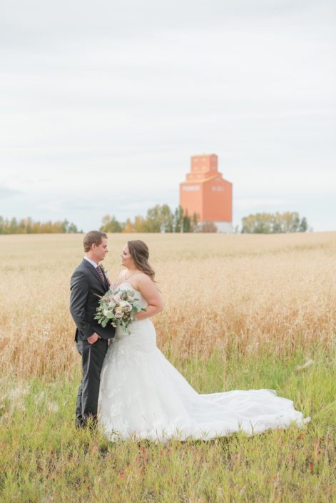 Bride and groom, Amber and Mike, beside a wheat field and grain elevator with mauve and ivory bridal bouquet featuring panda anemones, quicksand and amnesia roses and eucalyptus.