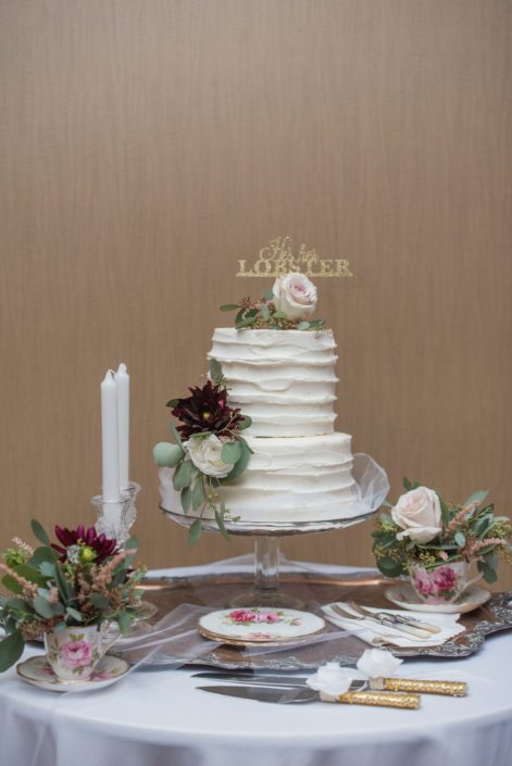 White wedding cake with cake flowers designed with quicksand roses, burgundy dahlias, and ranunculus accented by greenery with a gold "he's her lobster" cake topper.
