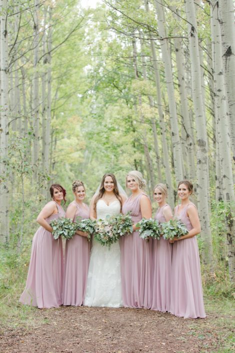 Bride, Amber, holding mauve and ivory bridal bouquet featuring roses and panda anemones with her bridesmaids wearing mauve floor length dresses and holding entirely greenery bouquets.