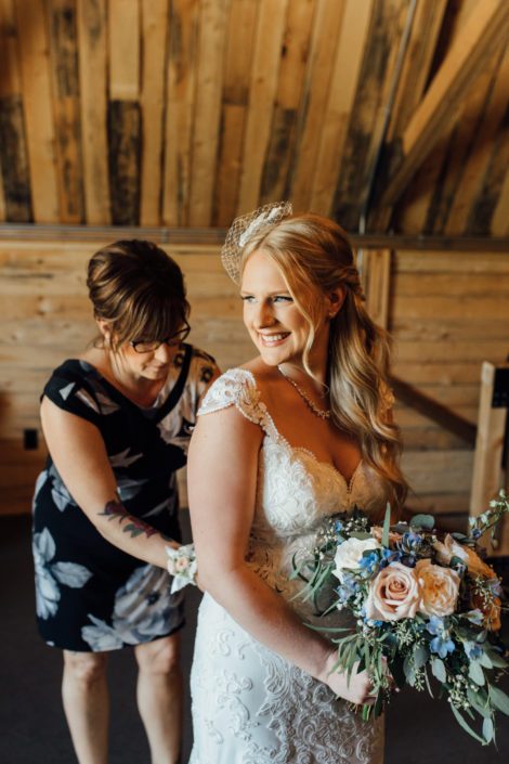 Kelsie and her mom doing up her bridal gown and holding her blush and blue bridal bouquet featuring white o'hara garden roses, quicksand roses, delphinium, gypsophila, and a mixed variety of eucalyptus greenery.