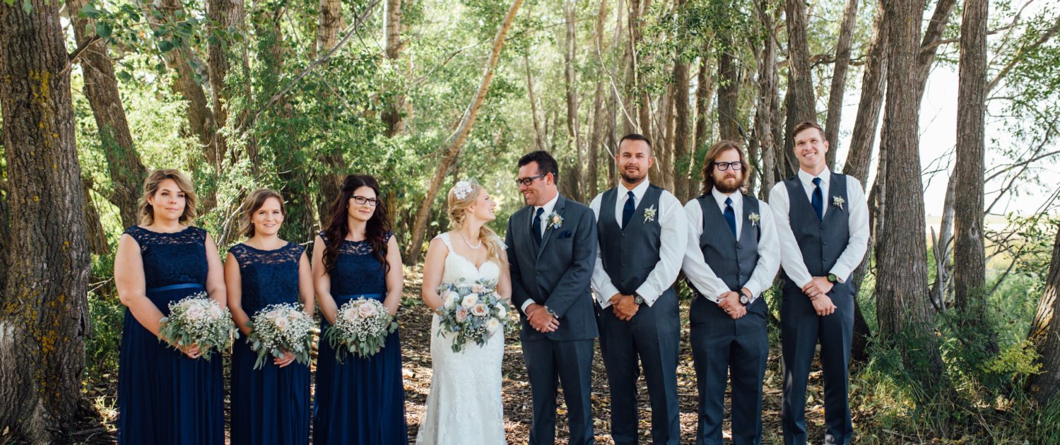 Kelsie and Kyle's bridal party; bridesmaids wearing blue dresses and holding bouquets designed with roses and babies breath; bride holding blush and blue bouquet designed with roses, delphinium, succulents, and babies breath; groom and groomsmen wearing boutonnieres.