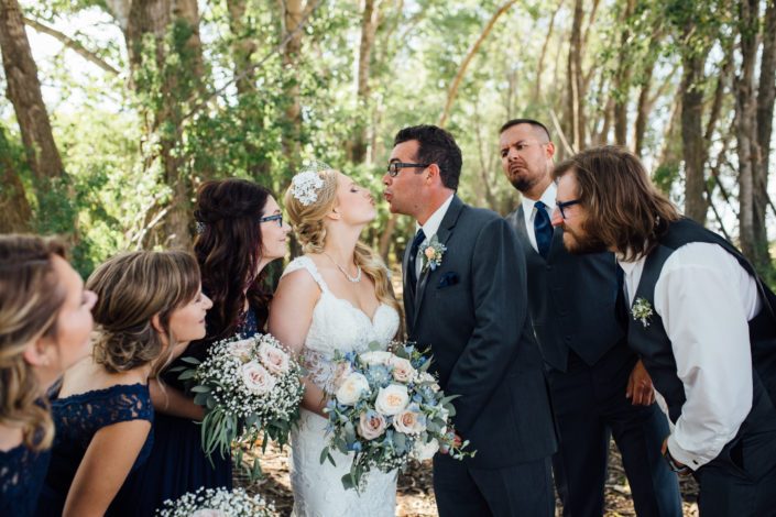 Bridal party watching as bride and groom are about to kiss; bridal bouquet featuring blue delphiniums, succulents, quicksand roses, white o'hara garden roses, babies breath and eucalyptus greenery.