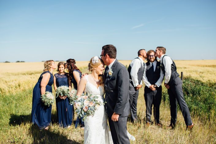 Bridal party being goofy by a field of wheat holding bouquets designed with roses, succulents, blue delphiniums, babies breath and eucalyptus.