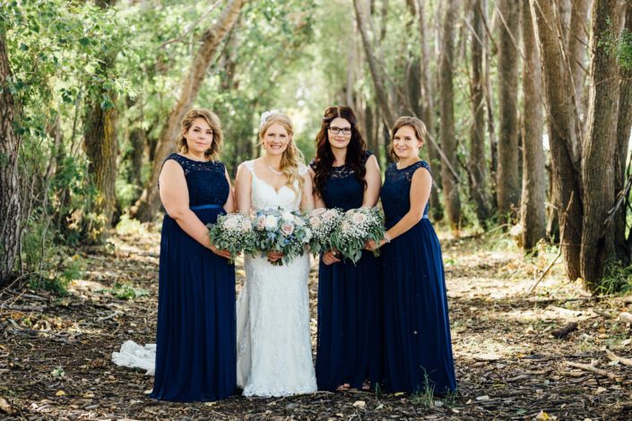 Bride and her bridesmaids holding bouquets featuring quicksand roses, white o'hara garden roses, succulents, delphiniums, babies breath and a mixed variety of eucalyptus greenery; bride wearing vintage inspired birdcage veil; bridesmaids wearing dark blue floor-length dresses.