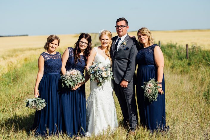 Bride and groom with bridesmaids near a field of wheat with bouquets made of blush roses, blue delphiniums, succulents, babies breath and eucalyptus greenery.