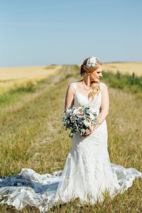 Bride standing by a field of wheat holding a blush and blue bridal bouquet featuring quicksand roses, white o'hara garden roses, blue delphinium, gypsophila (babies breath) and a mixed variety of eucalyptus.