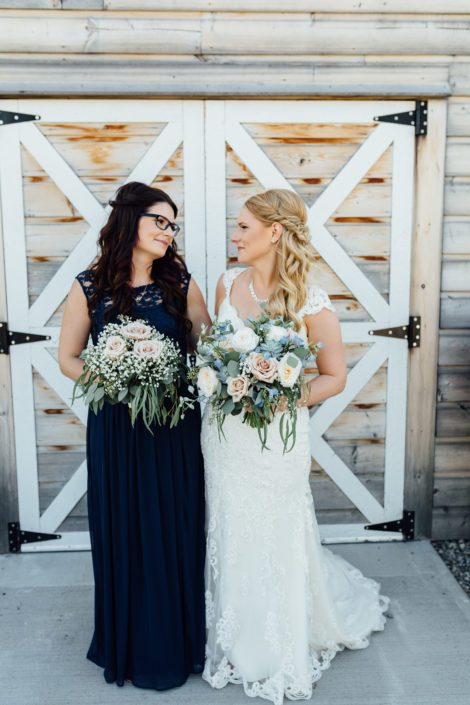 Bride and bridesmaid beside barn doors holding bouquets made of roses and babies breath with eucalyptus greenery; bridal bouquet also features succulents and blue delphiniums.