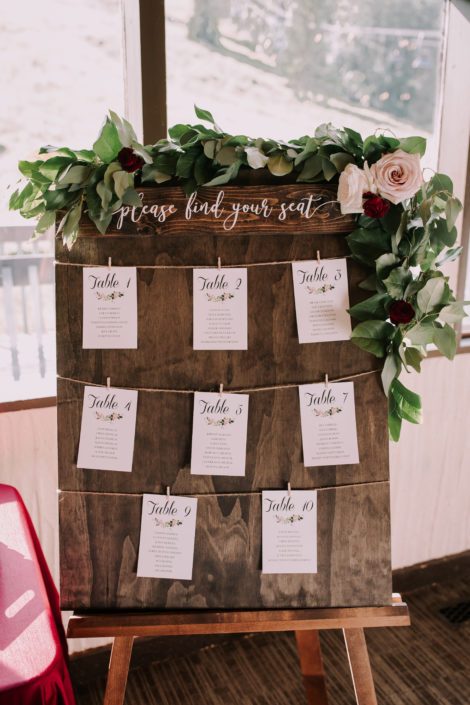 Seating chart sign with fresh greenery garland accented by burgundy and blush flowers.